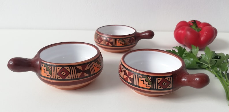 May include: Three small terracotta bowls with white interiors and brown handles. The bowls are decorated with geometric patterns in orange, brown, green, and red.