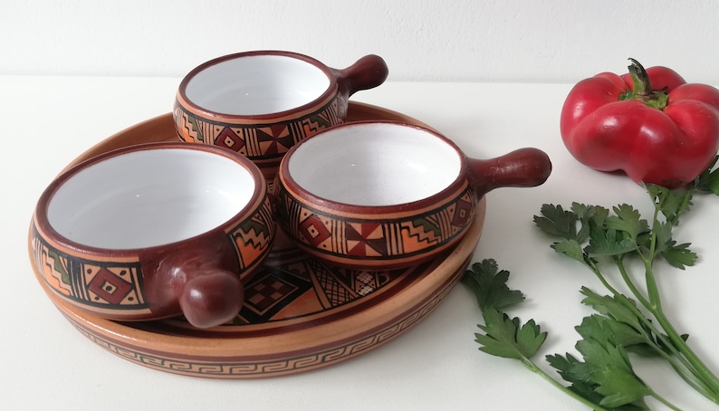May include: Set of three small terracotta bowls with white interiors and brown handles. The bowls are decorated with geometric patterns in shades of brown, orange, and green. The bowls are arranged on a larger terracotta platter with a similar geometric pattern.