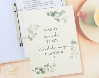 May include: A white wedding planner with a floral design and the text "MADDY and TOM'S Wedding PLANNER".