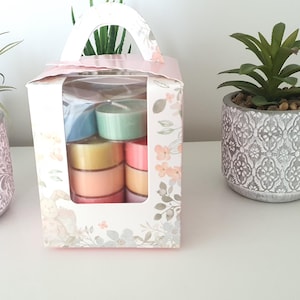 Könnte beinhalten: Eine Geschenkbox mit Griff, die bunte Teelichter enthält. Die Box hat ein Blumenmuster und ein klares Fenster. Zwei dekorative Sukkulenten in Töpfen befinden sich auf beiden Seiten der Box. Ein goldener Mosaik-Kerzenhalter befindet sich rechts.