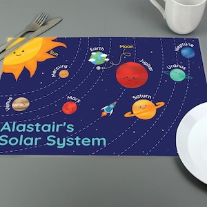 May include: A blue placemat with a colourful illustration of the solar system. The placemat features the sun, planets, and a rocket ship. The text "Alastair's Solar System" is printed on the placemat.