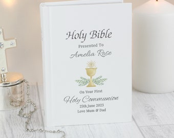 May include: White Holy Bible book with gold lettering and a watercolor illustration of a chalice with a host. The text reads "Holy Bible", "Presented To Amelia Rose", "On Your First Holy Communion", "25th June 2023", and "Love Mum & Dad".