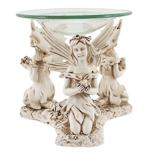 May include: A decorative oil warmer featuring three kneeling fairy figurines supporting a clear glass dish. The fairies are sculpted in a cream-colored resin with detailed wings and floral accents. The glass dish is round and has a green tint.