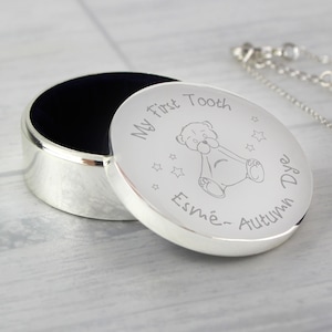 Könnte beinhalten: Eine silberfarbene, ovale Andenkenbox mit leicht geöffnetem Deckel. Der Deckel ist mit den Worten "My first Tooth", einer Teddybär-Illustration und dem Namen "Esme-Autumn Dye" graviert. Der Innenraum ist mit dunkelblauem Samt ausgekleidet.