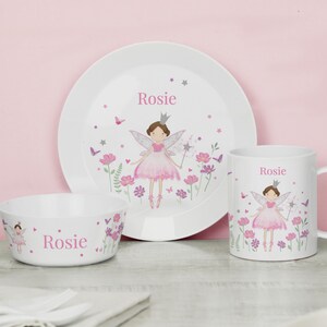 May include: A personalised children's dinnerware set featuring a pink fairy with wings and a crown. The set includes a bowl, plate and mug with the name "Rosie" printed on each piece. The design includes pink flowers, butterflies and stars on a white background.