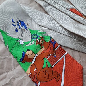 May include: A brown and orange tie with a cartoon image of a bear playing tennis. The bear is wearing a green hat and holding a tennis racket. The background is a green tennis court with a white net. The tie has a white and gray pattern on the back.