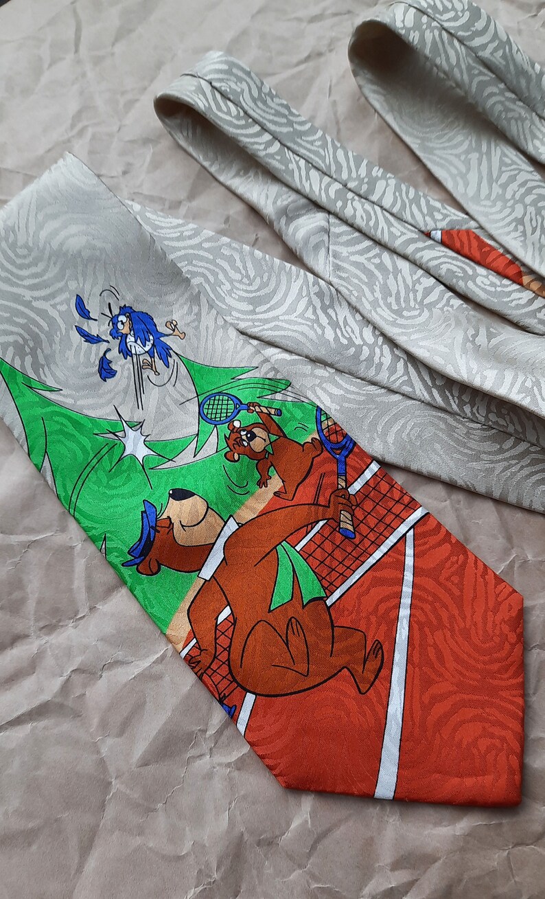 May include: A brown and orange tie with a cartoon image of a bear playing tennis. The bear is wearing a green hat and holding a tennis racket. The background is a green tennis court with a white net. The tie has a white and gray pattern on the back.