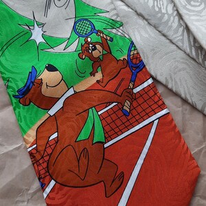 May include: A red, green, and white tie with a cartoon image of two bears playing tennis. The bear on the left is holding a tennis racket and the bear on the right is running toward the net. A blue bird is flying above the bears. The tie has a white and orange pattern.