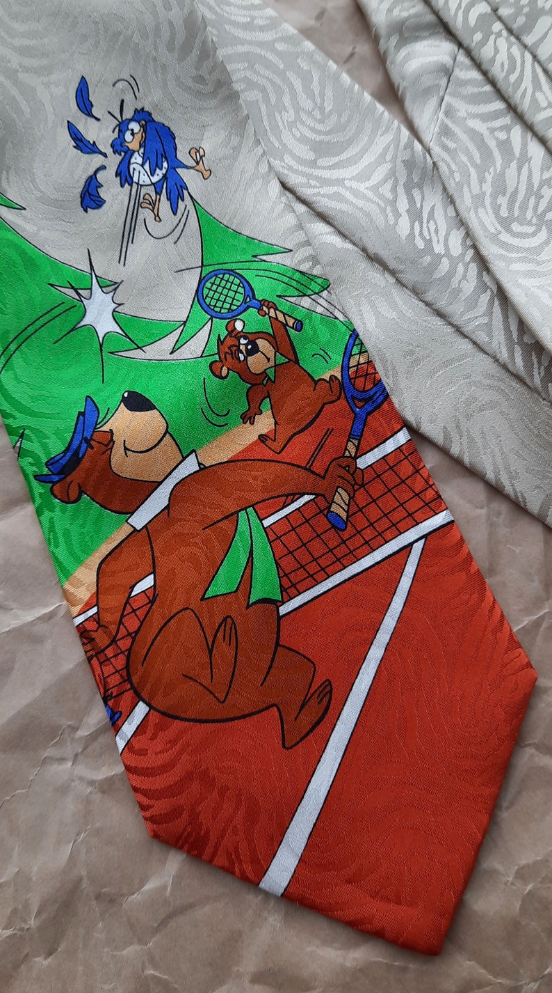May include: A red, green, and white tie with a cartoon image of two bears playing tennis. The bear on the left is holding a tennis racket and the bear on the right is running toward the net. A blue bird is flying above the bears. The tie has a white and orange pattern.