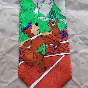 May include: A brown bear wearing a blue hat and green scarf is playing tennis with a smaller brown bear. The bears are on a red and white tennis court with a green tree in the background. A blue bird with white feathers is flying in the air above the bears. The tie is made of silk and has a shiny finish.