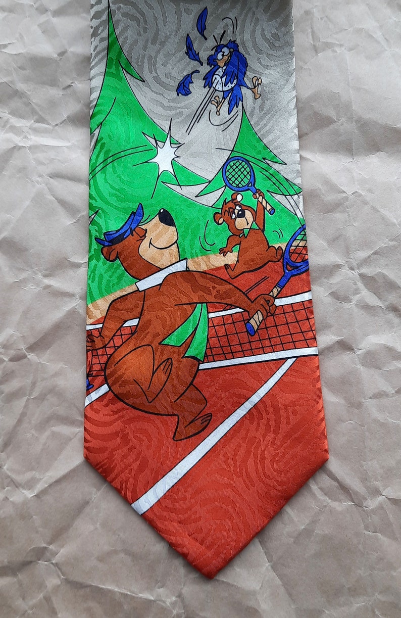 May include: A brown bear wearing a blue hat and green scarf is playing tennis with a smaller brown bear. The bears are on a red and white tennis court with a green tree in the background. A blue bird with white feathers is flying in the air above the bears. The tie is made of silk and has a shiny finish.