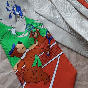 May include: A silk tie with a green, orange, and white tennis court design. The tie features a cartoon bear with a blue hat holding a tennis racket and a blue bird flying away from the bear. The tie has a white background with a subtle gray pattern.