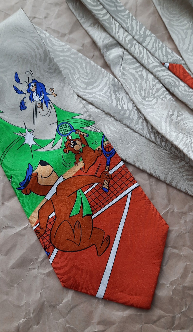 May include: A silk tie with a green, orange, and white tennis court design. The tie features a cartoon bear with a blue hat holding a tennis racket and a blue bird flying away from the bear. The tie has a white background with a subtle gray pattern.