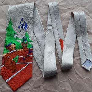 May include: A tie with a white background and a repeating pattern of swirls. The tie features a cartoon image of two bears playing tennis with a bird watching from above. The tennis court is red and orange with white lines. The bears are brown and wearing blue hats. The bird is blue with white wings. The tie is tied in a knot and the ends are hanging down.