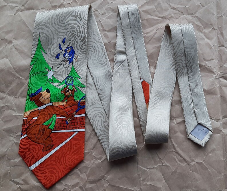 May include: A tie with a white background and a repeating pattern of swirls. The tie features a cartoon image of two bears playing tennis with a bird watching from above. The tennis court is red and orange with white lines. The bears are brown and wearing blue hats. The bird is blue with white wings. The tie is tied in a knot and the ends are hanging down.