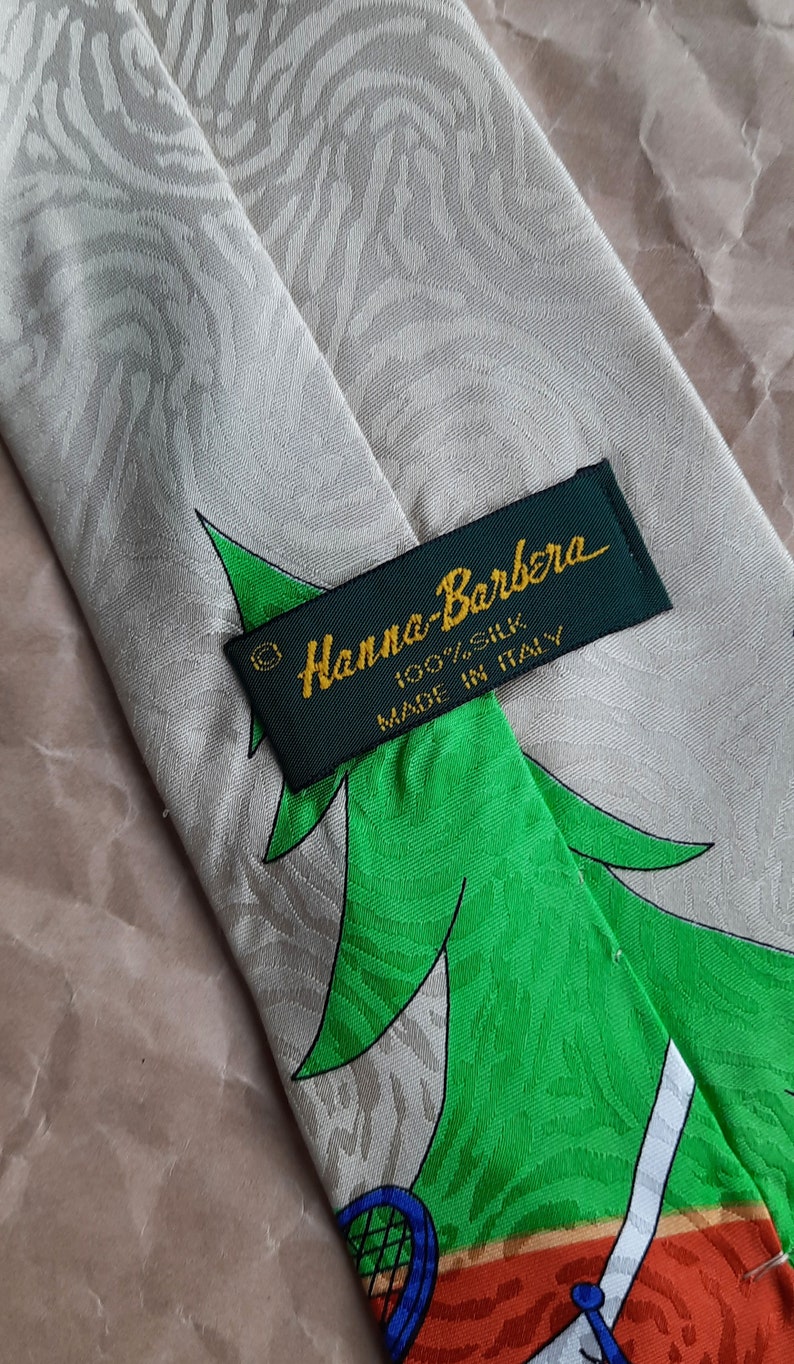 May include: A green, orange, and blue patterned silk tie with a green label that reads 'Hanna-Barbera 100% Silk Made in Italy'.