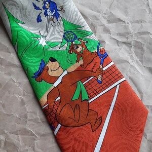 May include: A brown and green tie with a cartoon image of Yogi Bear and Boo Boo Bear playing tennis. The background is a green and white striped pattern with a tennis net and a red clay tennis court.
