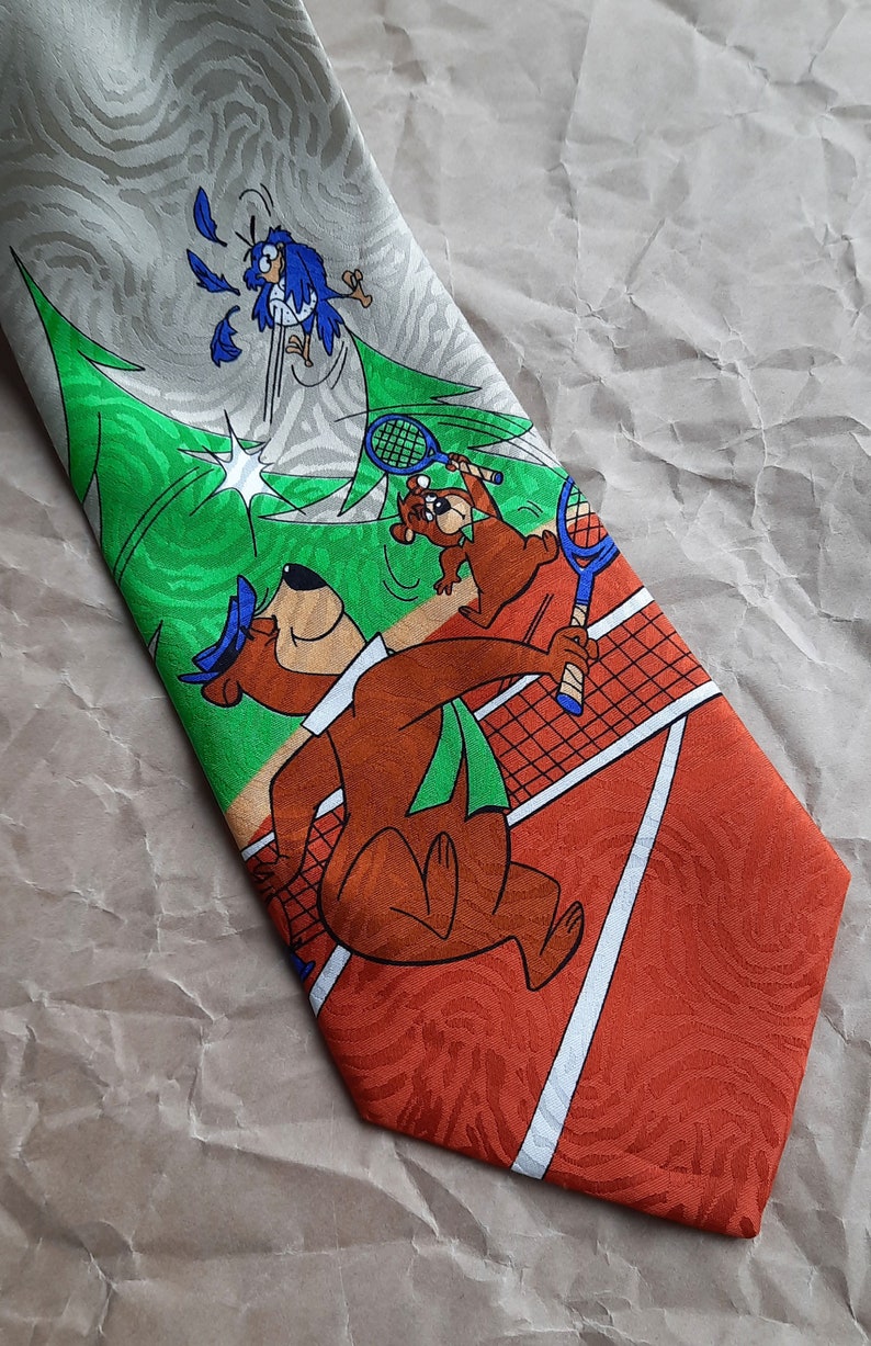 May include: A brown and green tie with a cartoon image of Yogi Bear and Boo Boo Bear playing tennis. The background is a green and white striped pattern with a tennis net and a red clay tennis court.