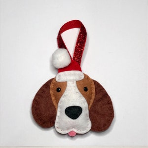 May include: A felt Christmas ornament shaped like a dog wearing a Santa hat with a white pom-pom. The dog has brown ears, a white face, and a pink tongue.