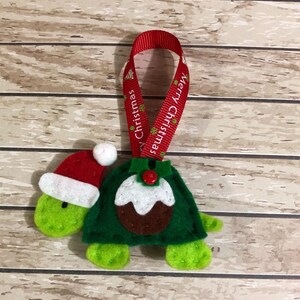 May include: Green felt Christmas ornament shaped like a turtle wearing a Santa hat and a green felt Christmas pudding. The ornament has a red ribbon with the text "Merry Christmas" printed on it.