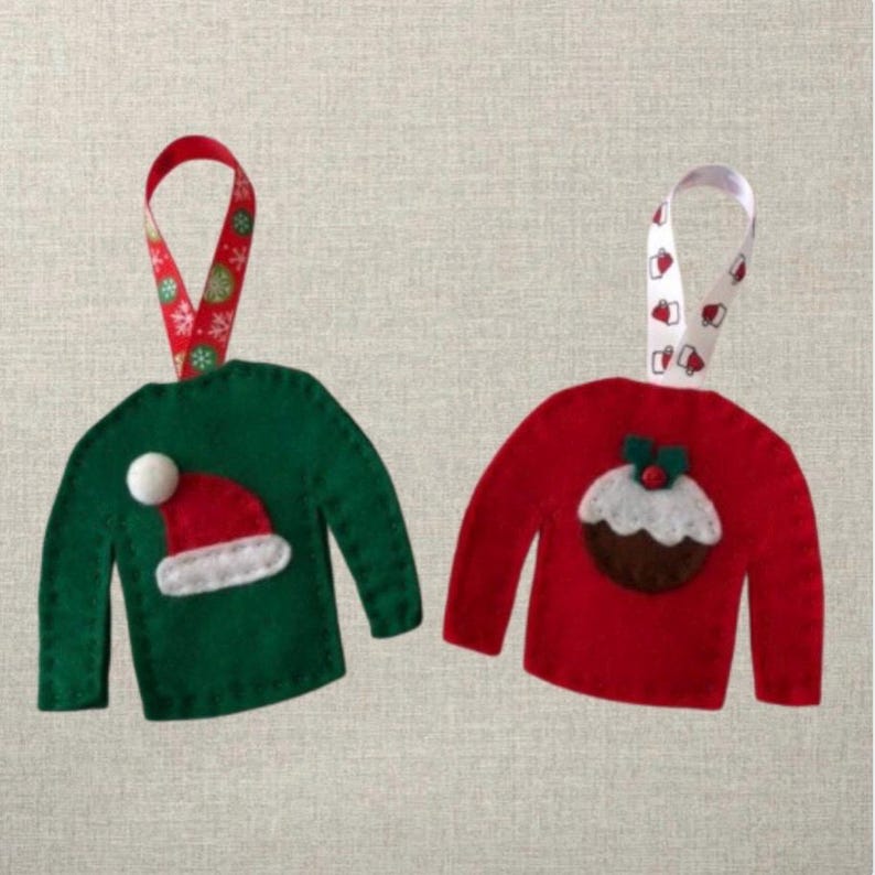 May include: Two felt Christmas ornaments shaped like sweaters. One sweater is green with a white pom-pom and a red Santa hat. The other sweater is red with a brown and white Christmas pudding.