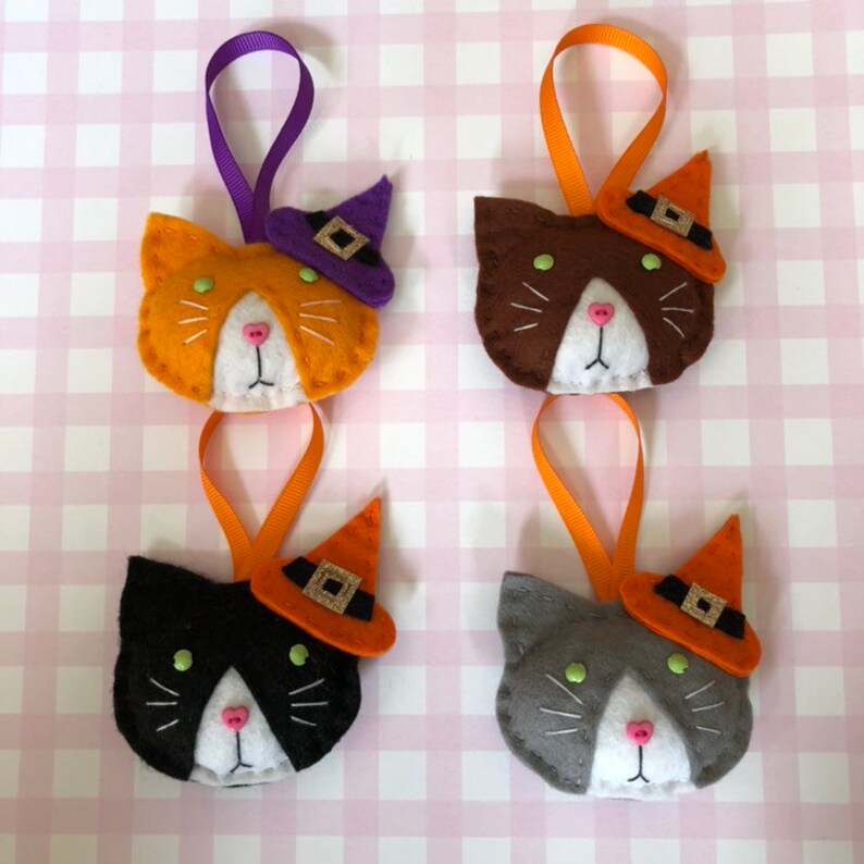 May include: Four felt cat ornaments in orange, brown, black, and gray, each with a witch hat and ribbon loop. The cats have white faces, pink noses, and green eyes. The hats are orange with black bands.