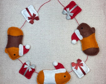 Peut inclure: Une guirlande d'ornements en feutre représentant des cochons d'Inde bruns et orange portant des bonnets de Père Noël et des boîtes cadeaux rouges et blanches avec des nœuds argentés.