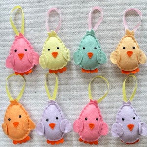 May include: Eight felt bird ornaments in various pastel colors, including pink, yellow, light blue, lavender, and orange. Each bird has a small beak and black eyes. The birds are hanging from yellow and pink ribbons.