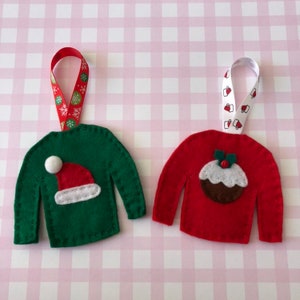 May include: Two felt Christmas ornaments shaped like sweaters. One is green with a white Santa hat and the other is red with a brown Christmas pudding.
