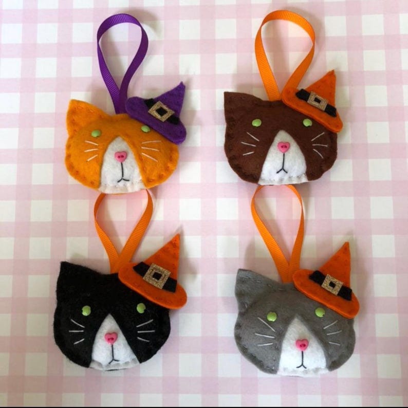 May include: Four felt cat ornaments in orange, brown, black, and gray, each with a white face, pink nose, and green eyes. Each cat wears a witch hat and has a ribbon for hanging. The background is a pink and white checkered pattern.