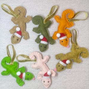 Peut inclure: Ornements de lézard en feutre de différentes couleurs, notamment beige, vert, orange et rose. Chaque lézard porte un petit bonnet de Père Noël et un ruban doré pour l'accrocher. Ces décorations sont pour Noël.