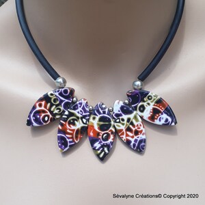 Puede incluir: Un collar gargantilla negro con cinco colgantes en forma de hoja coloridos y con patrones. Los colgantes están hechos de arcilla polimérica y tienen una combinación de colores negro, blanco, morado, naranja y verde.