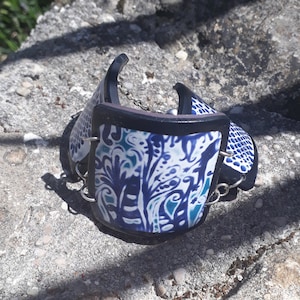 May include: A blue and white patterned bracelet with a black leather band and silver chain. The bracelet is made of polymer clay and has a unique design.