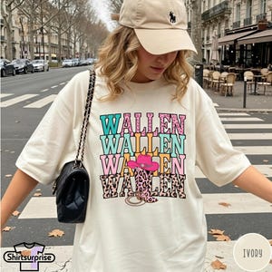 Op de afbeelding: Een ivoorkleurig T-shirt met korte mouwen en de tekst WALLEN vier keer herhaald in turquoise, roze en luipaardprint. Een roze cowboyhoed en een cowboylaars met luipaardprint zijn in het ontwerp verwerkt. Een zwarte schoudertas wordt gedragen.