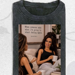 Peut inclure: Un t-shirt gris anthracite délavé avec une impression graphique représentant deux poupées assises sur un canapé. L'une des poupées tient une tasse blanche et fait une grimace de dégoût. Au-dessus, un panneau indique : When someone asks when I'm going to start dating again.