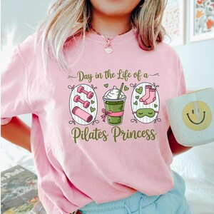 K&ouml;nnte beinhalten: Hellrosa T-Shirt mit dem Text "Day in the Life of a Pilates Princess" und Illustrationen von Hanteln, einem Getr&auml;nk, Socken und einer Schlafmaske. Eine wei&szlig;e Tasse mit einem Smiley wird gehalten.