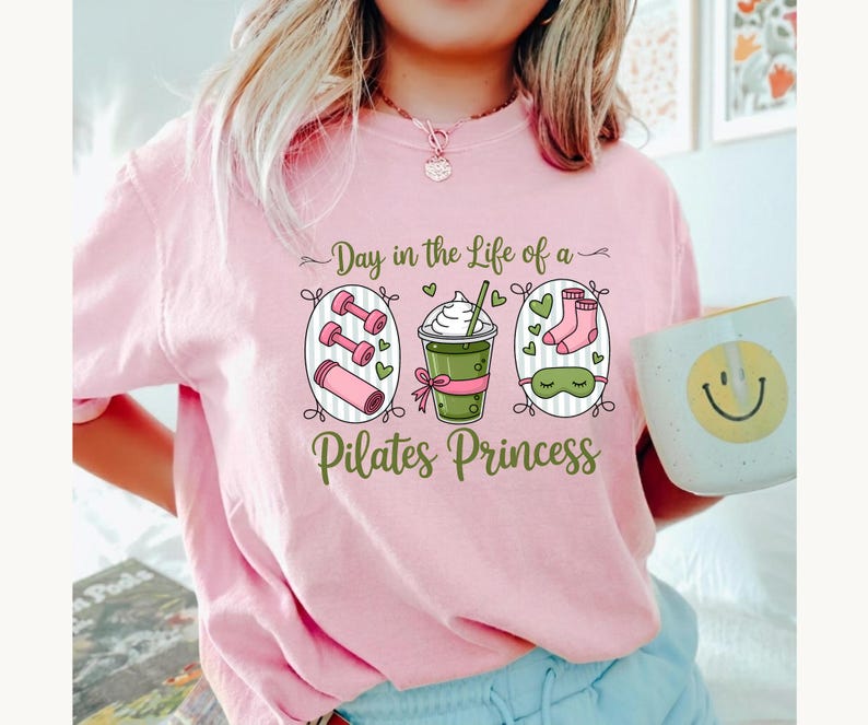 K&ouml;nnte beinhalten: Hellrosa T-Shirt mit dem Text "Day in the Life of a Pilates Princess" und Illustrationen von Hanteln, einem Getr&auml;nk, Socken und einer Schlafmaske. Eine wei&szlig;e Tasse mit einem Smiley wird gehalten.