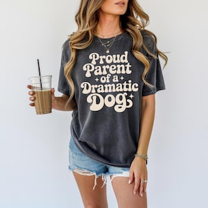 May include: Dark gray t-shirt with the text "Proud Parent of a Dramatic Dog" in a retro font. The shirt is a relaxed fit and has a vintage wash. The shirt is paired with denim shorts.