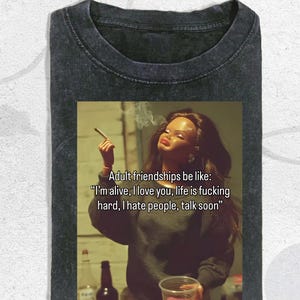 May include: A charcoal grey acid-wash t-shirt featuring a graphic print of a doll with long dark hair holding a cigarette. The text reads: Adult friendships be like: I’m alive, I love you, life is fucking hard, I hate people, talk soon.