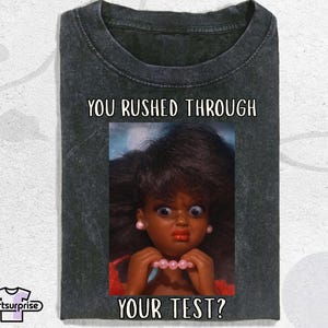 May include: Dark grey t-shirt featuring the text "YOU RUSHED THROUGH YOUR TEST?" above and below an image of a doll. The doll has a surprised expression, wearing a pink necklace and earrings.