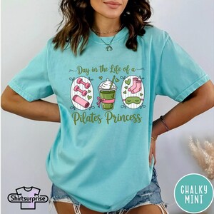 K&ouml;nnte beinhalten: Hellblaues T-Shirt mit dem Aufdruck "Day in the Life of a Pilates Princess". Die Grafik zeigt Hanteln, ein Getr&auml;nk, Socken und eine Schlafmaske. Das Shirt hat kurze &Auml;rmel und eine lockere Passform.