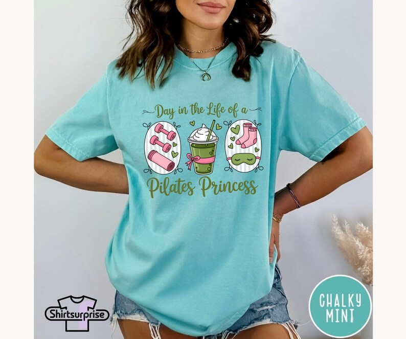 K&ouml;nnte beinhalten: Hellblaues T-Shirt mit dem Aufdruck "Day in the Life of a Pilates Princess". Die Grafik zeigt Hanteln, ein Getr&auml;nk, Socken und eine Schlafmaske. Das Shirt hat kurze &Auml;rmel und eine lockere Passform.