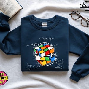 May include: Navy blue sweatshirt featuring a colorful Rubik's Cube design with mathematical equations. The graphic includes a detailed illustration of the cube and various formulas. The sweatshirt has a crew neck and long sleeves.