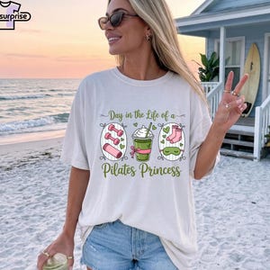 K&ouml;nnte beinhalten: Wei&szlig;es T-Shirt mit dem Text "Day in the Life of a Pilates Princess". Die Grafik zeigt Hanteln, einen Smoothie und Socken. Das Shirt hat eine lockere Passform.
