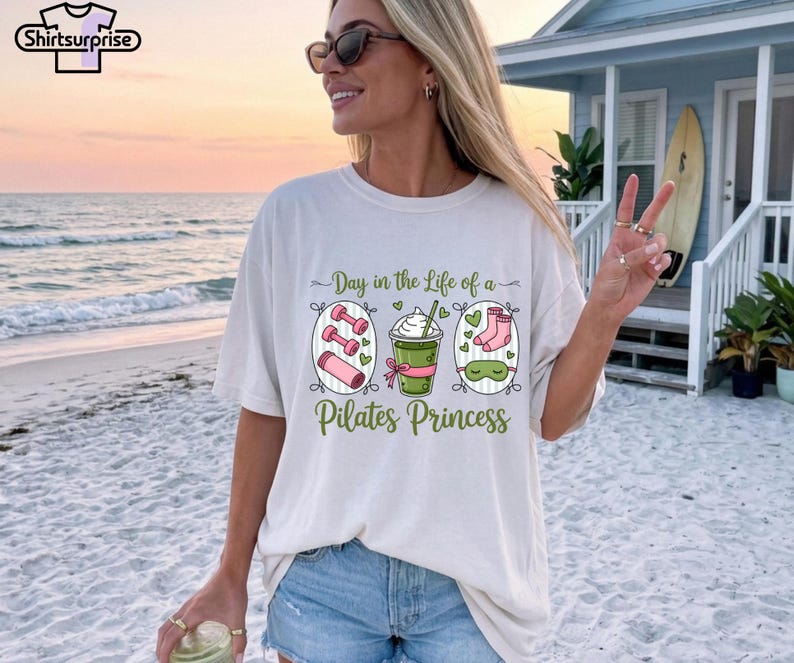 K&ouml;nnte beinhalten: Wei&szlig;es T-Shirt mit dem Text "Day in the Life of a Pilates Princess". Die Grafik zeigt Hanteln, einen Smoothie und Socken. Das Shirt hat eine lockere Passform.