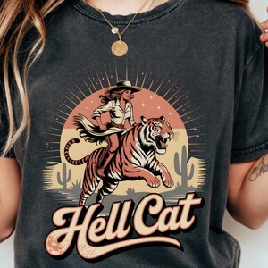 May include: Dark grey t-shirt with a retro design of a woman riding a tiger, set against a desert sunset and cacti. The words "Hell Cat" are in a vintage font. The graphic is printed on a soft, comfortable tee.