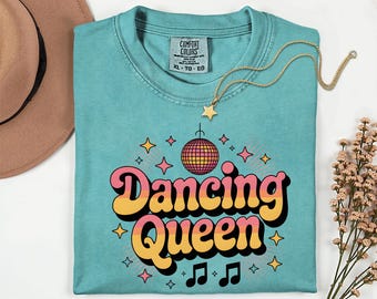 May include: A turquoise Comfort Colors t-shirt with the words "Dancing Queen" in a retro font, a disco ball graphic, and musical notes. A gold star pendant necklace rests on the shirt. A brown hat and flowers are in the background.