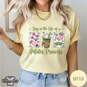K&ouml;nnte beinhalten: Hellgelbes T-Shirt mit dem Text "Day in the Life of a Pilates Princess". Das Design umfasst Illustrationen von Hanteln, einem Getr&auml;nk, Socken und einer Schlafmaske. Das T-Shirt hat kurze &Auml;rmel und eine lockere Passform.