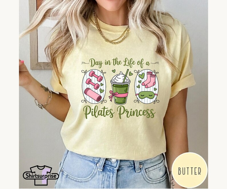 K&ouml;nnte beinhalten: Hellgelbes T-Shirt mit dem Text "Day in the Life of a Pilates Princess". Das Design umfasst Illustrationen von Hanteln, einem Getr&auml;nk, Socken und einer Schlafmaske. Das T-Shirt hat kurze &Auml;rmel und eine lockere Passform.