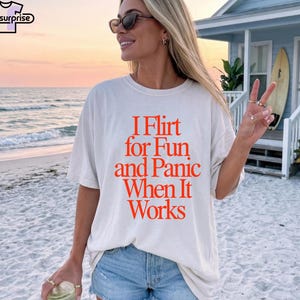 Könnte beinhalten: Cremefarbenes T-Shirt mit dem orangefarbenen Text "I Flirt for Fun and Panic When It Works". Das T-Shirt wird auf einem Strand getragen, mit einem Sonnenuntergang im Hintergrund.